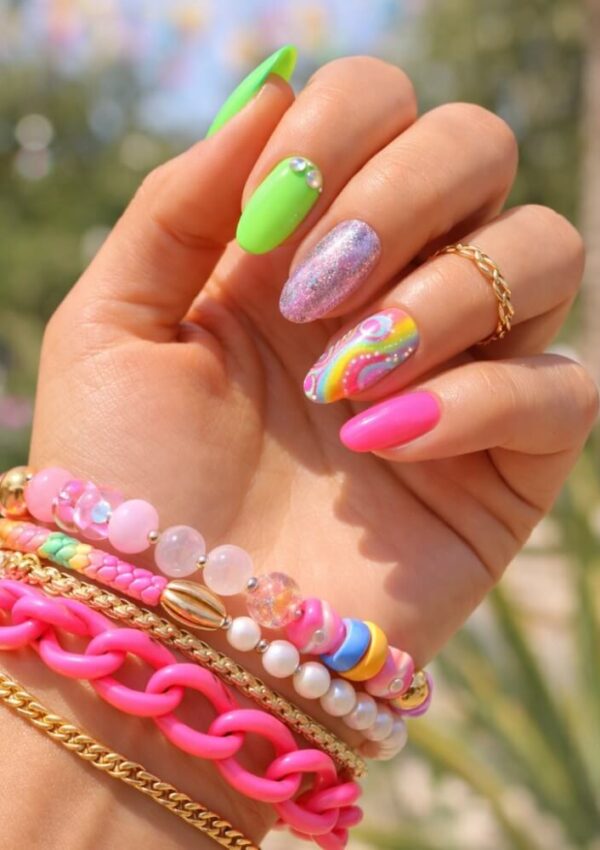 A hand with colorful manicured nails featuring neon green, hot pink, glitter, and a swirl design, adorned with beaded and gold bracelets and rings, held against a blurred outdoor background.