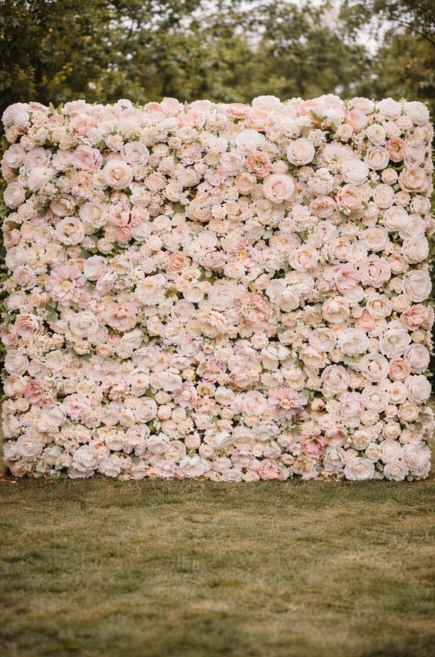 A large, square wall covered with an arrangement of pink, cream, and white roses and peonies stands on a grassy lawn with trees in the background.