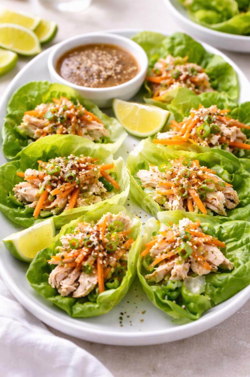 A plate of lettuce wraps filled with shredded chicken, julienned carrots, green onions, and sesame seeds, served with lime wedges and a bowl of dipping sauce on the side.