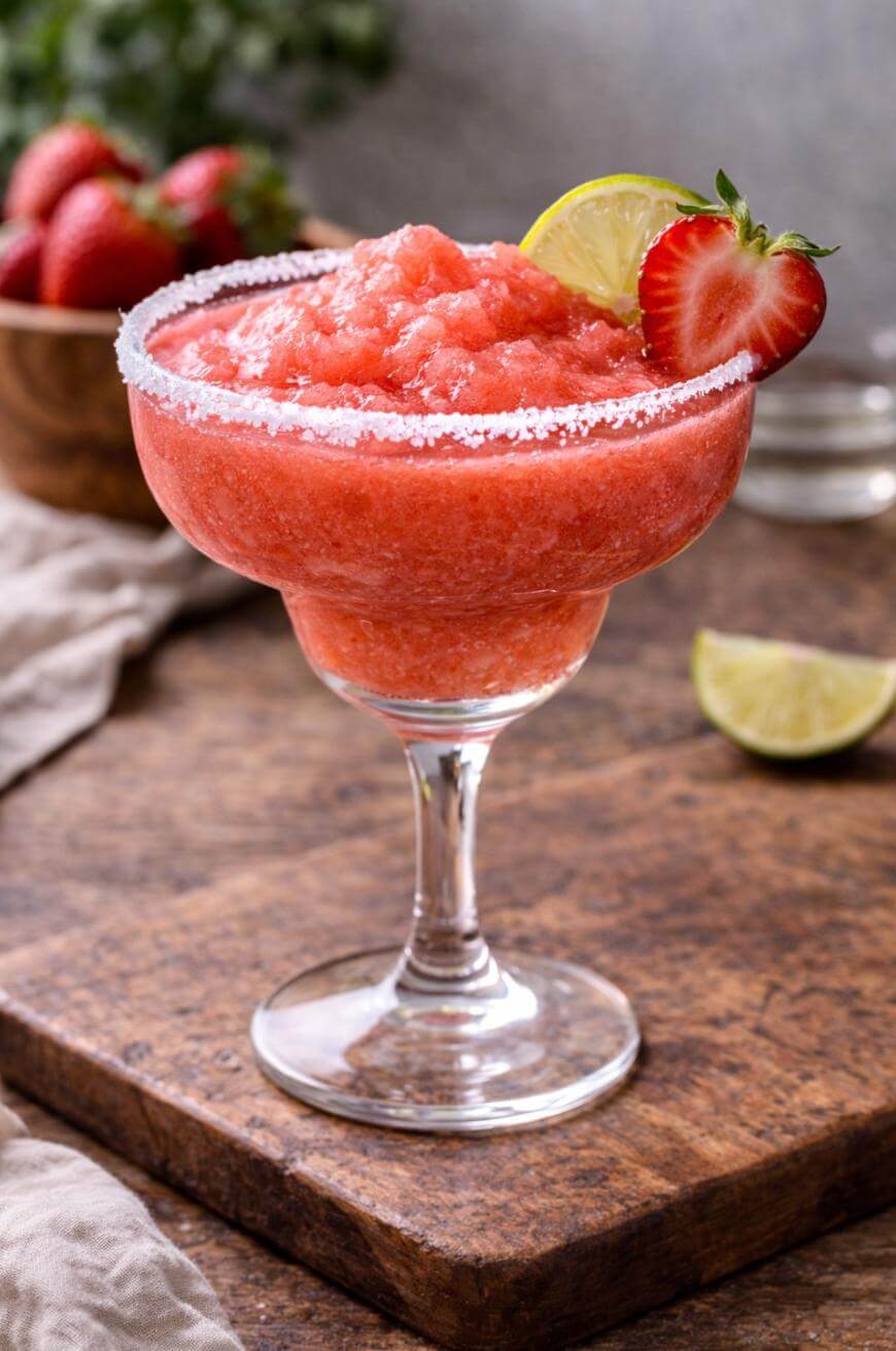 A strawberry frozen margarita in a glass with a salted rim, garnished with a fresh strawberry and a lime slice, sits on a wooden surface. A lime wedge and bowl of strawberries are in the background.