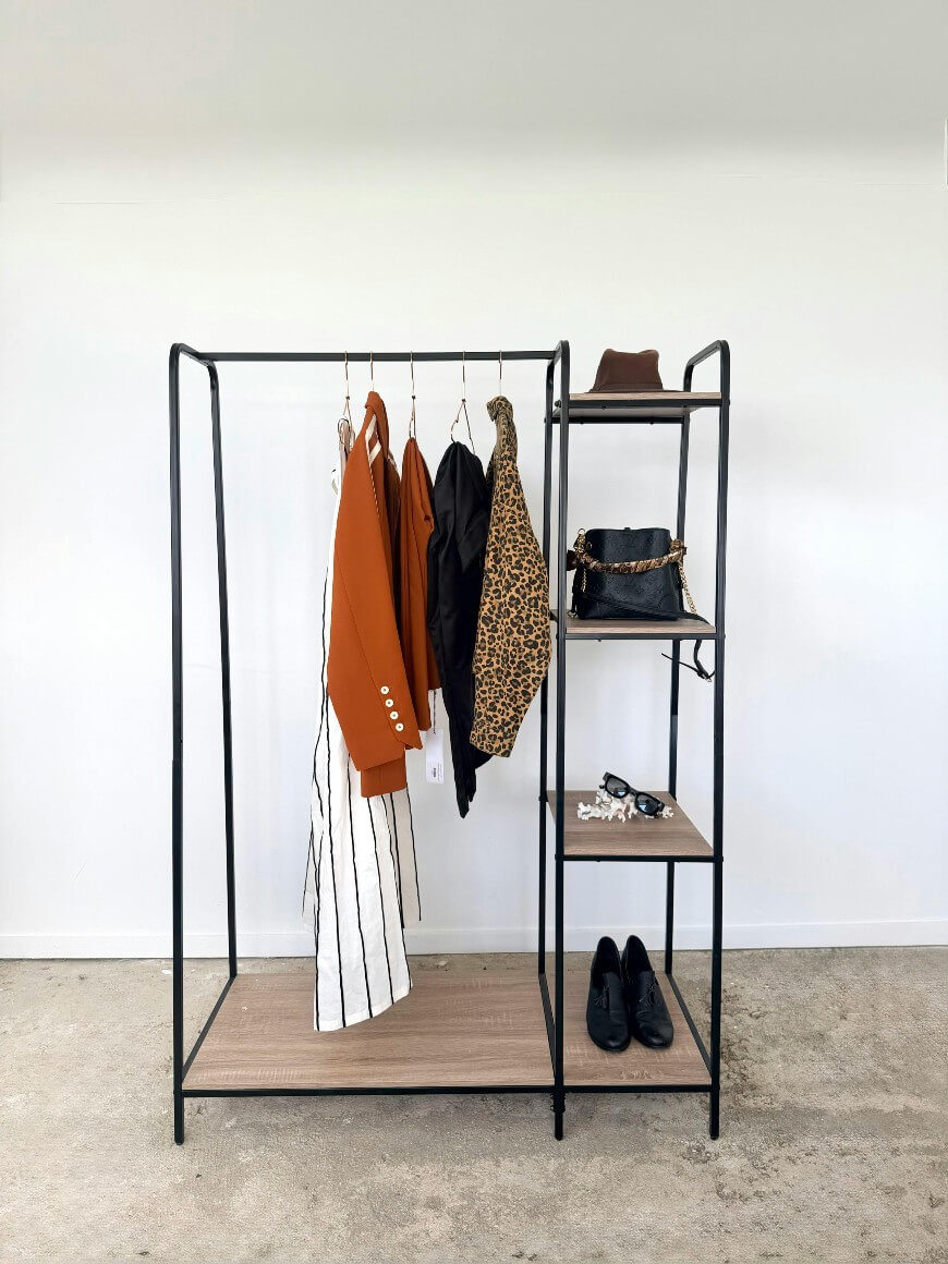 A black metal clothing rack with wood shelves displays four hanging garments, including a white striped dress, an orange jacket, a black jacket, and a leopard print coat. Shelves hold a hat, black handbag, jewelry, and black shoes.
