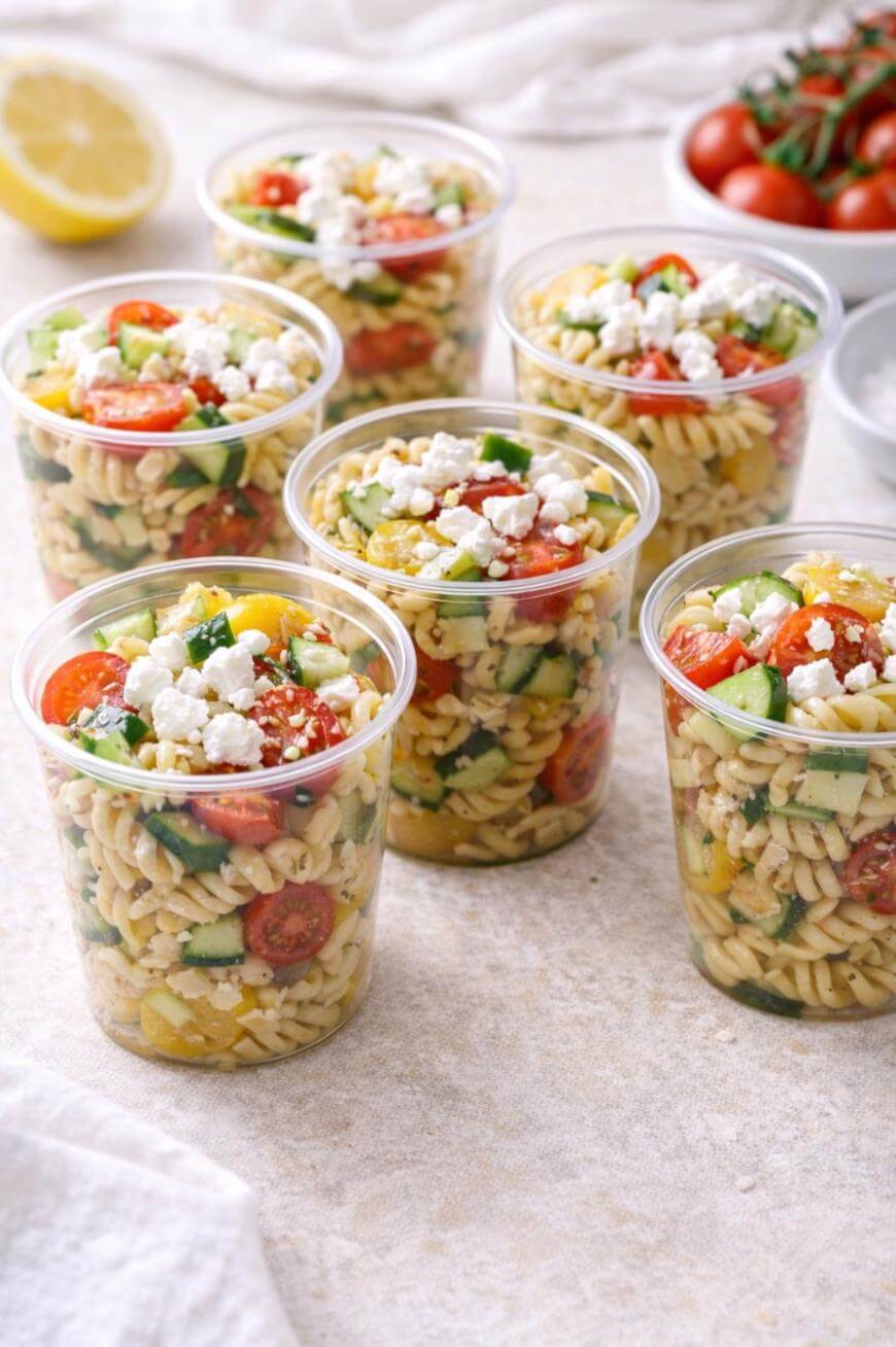 Six clear plastic cups filled with pasta salad made of rotini, cherry tomatoes, cucumbers, yellow bell peppers, and topped with crumbled feta cheese. A bowl of tomatoes and a lemon are in the background.