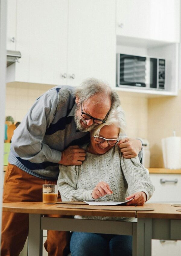 An elderly man hugs an elderly woman from behind as she smiles and uses a tablet at a kitchen table, with a glass of juice nearby and a microwave in the background.