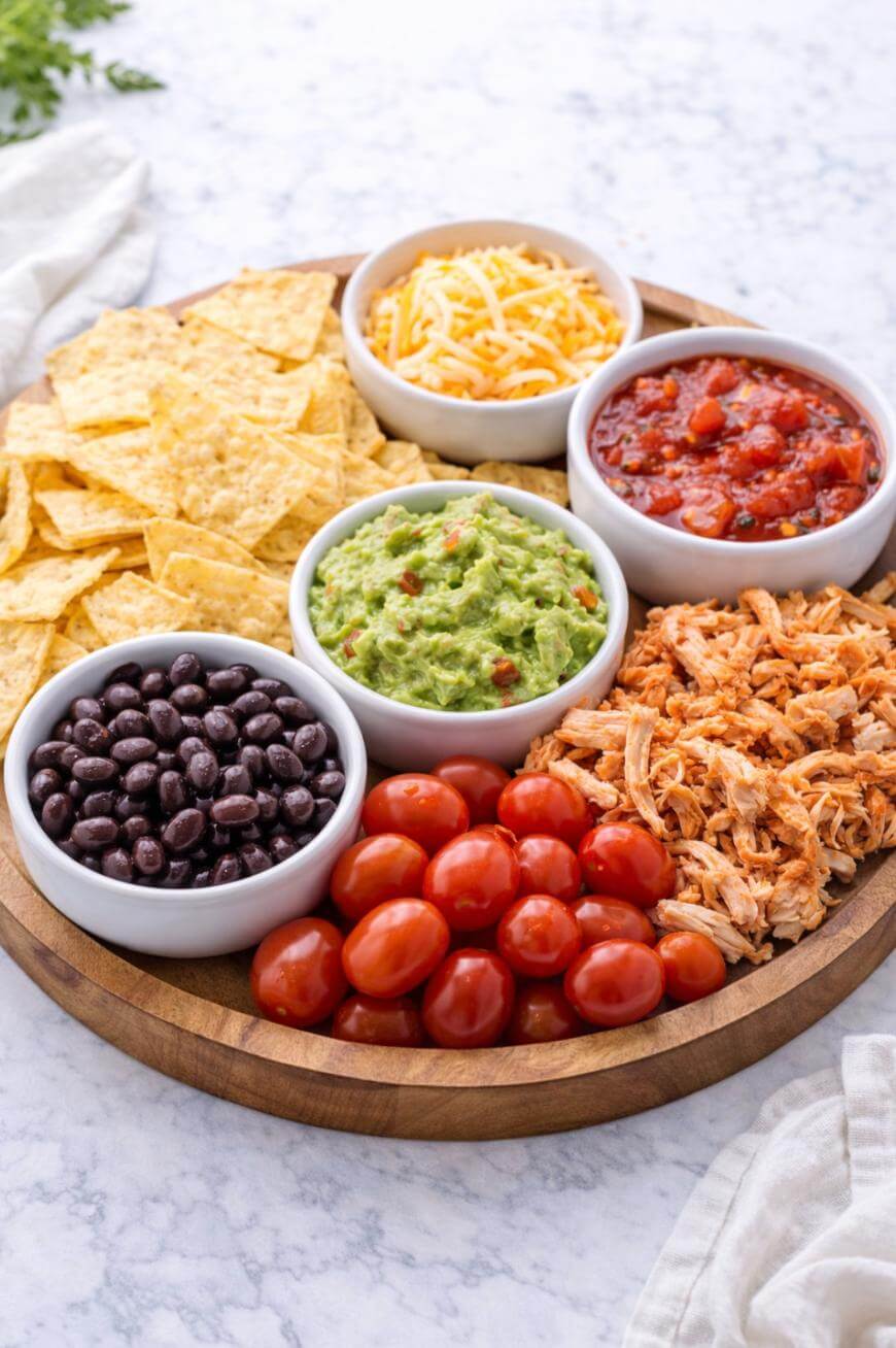 A round wooden platter with small bowls of shredded cheese, salsa, guacamole, black beans, tortilla chips, shredded chicken, and cherry tomatoes, arranged neatly on a marble surface.
