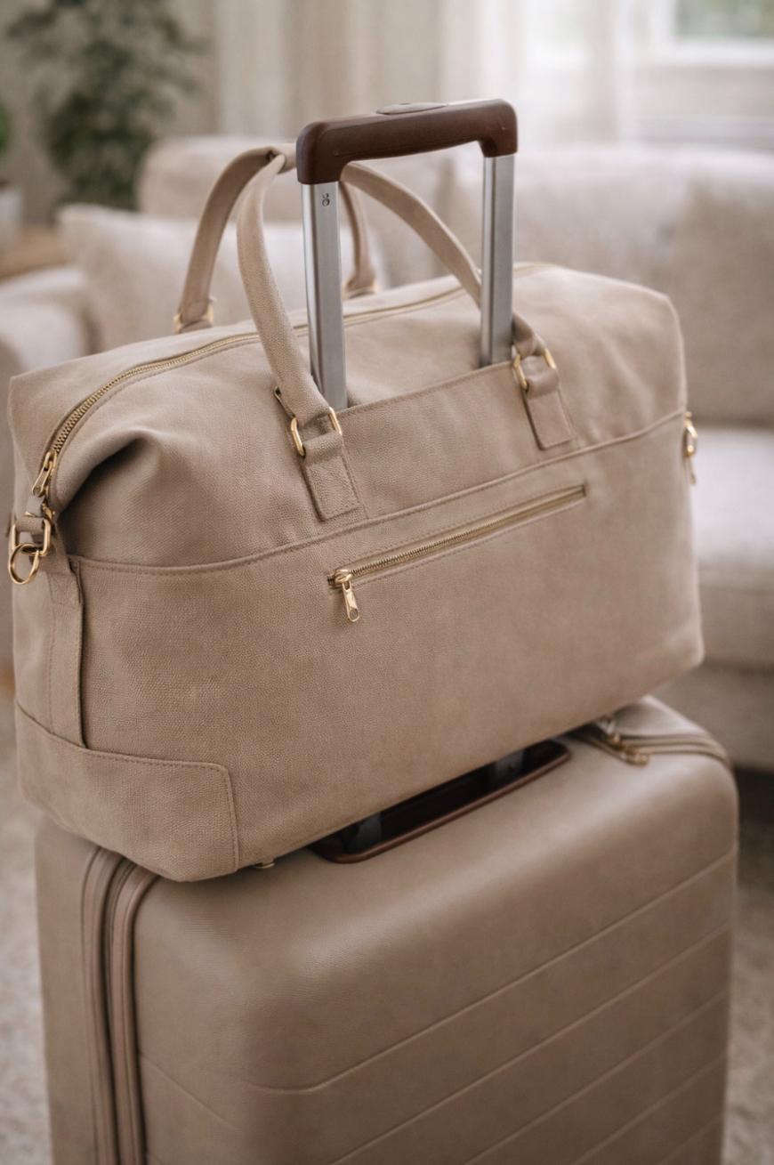 A tan duffel bag with gold zippers sits on top of a matching hard-shell suitcase with an extended handle, both placed indoors in a softly lit room with neutral decor.