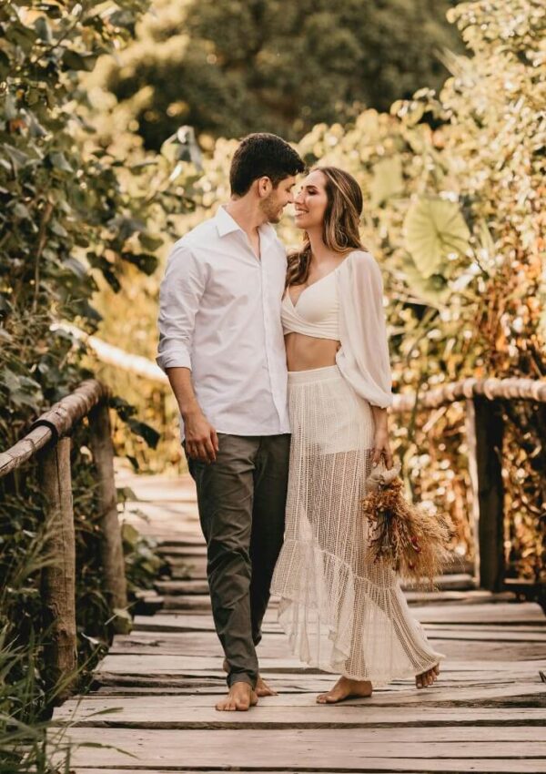 A couple walks barefoot on a wooden path surrounded by lush greenery. The woman wears a white two-piece outfit and holds a bouquet, while the man wears a white shirt and gray pants. They gaze at each other and smile.