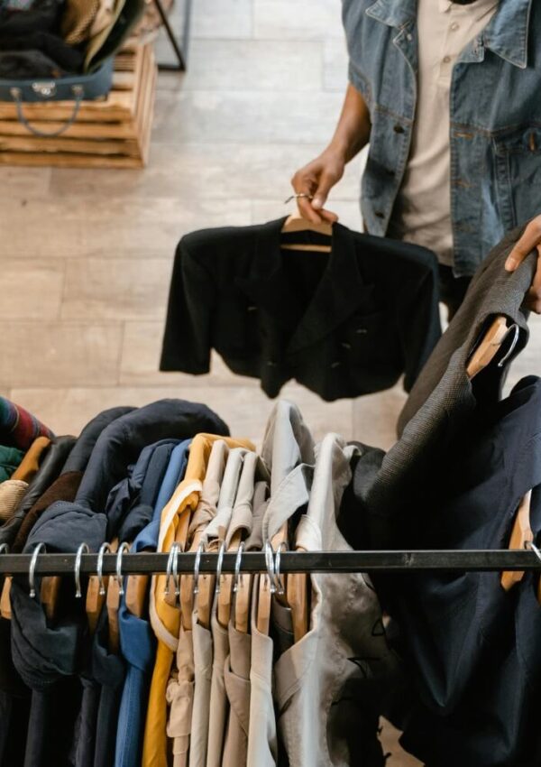 Person holding a black jacket on a hanger, browsing through a clothing rack filled with assorted jackets and shirts in a store with wooden floors.