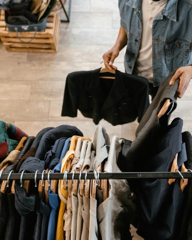 Person holding a black jacket on a hanger, browsing through a clothing rack filled with assorted jackets and shirts in a store with wooden floors.