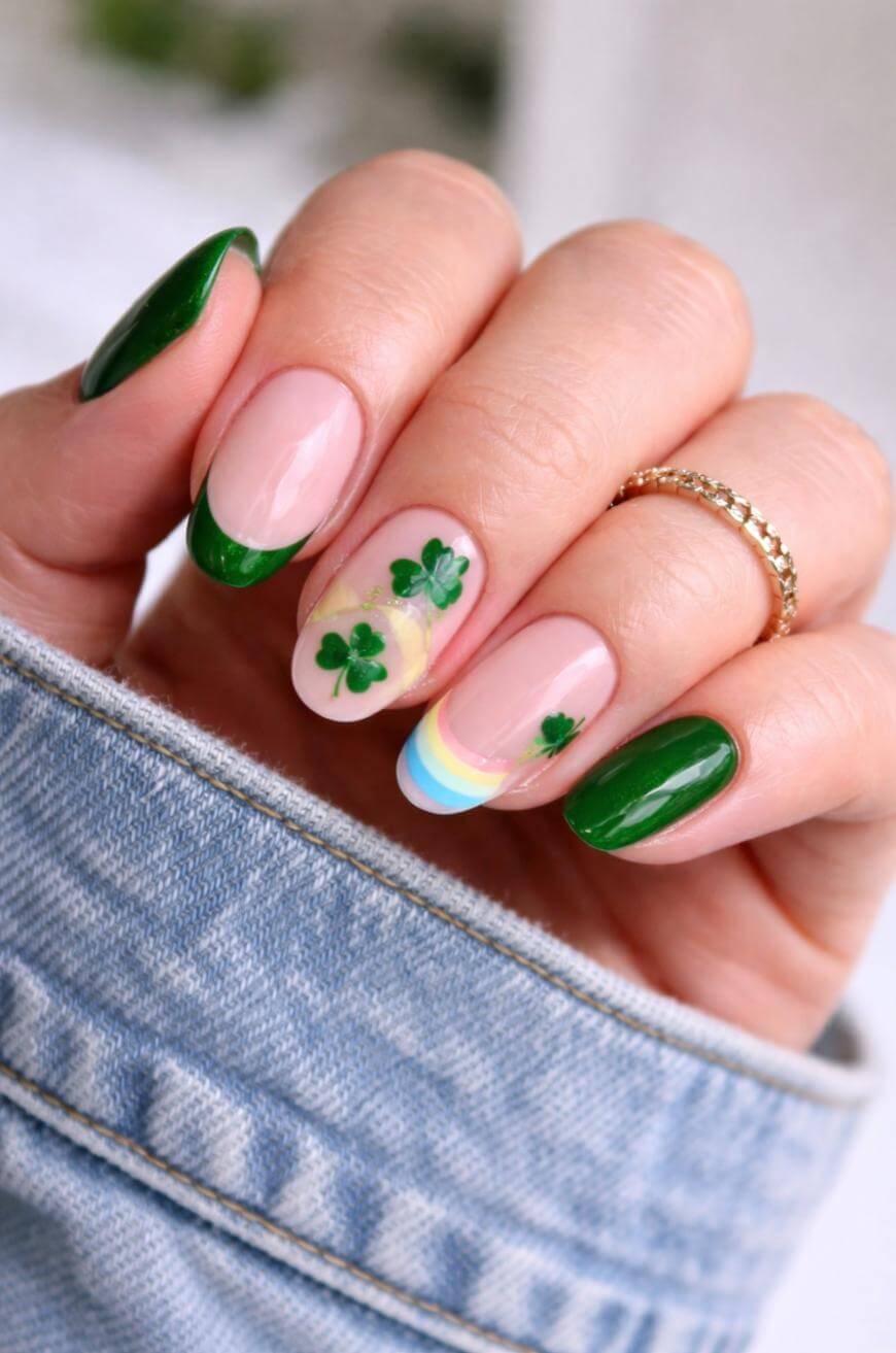 A hand with manicured nails featuring green tips, shamrock designs, and a small rainbow accent. The person is wearing a denim jacket and a gold ring.