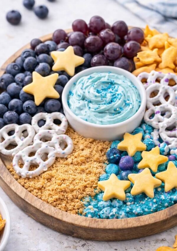 A round wooden tray with clusters of purple grapes, blueberries, yogurt-covered pretzels, goldfish crackers, star-shaped cookies, blue candies, cookie crumbs, and a bowl of blue dip in the center.
