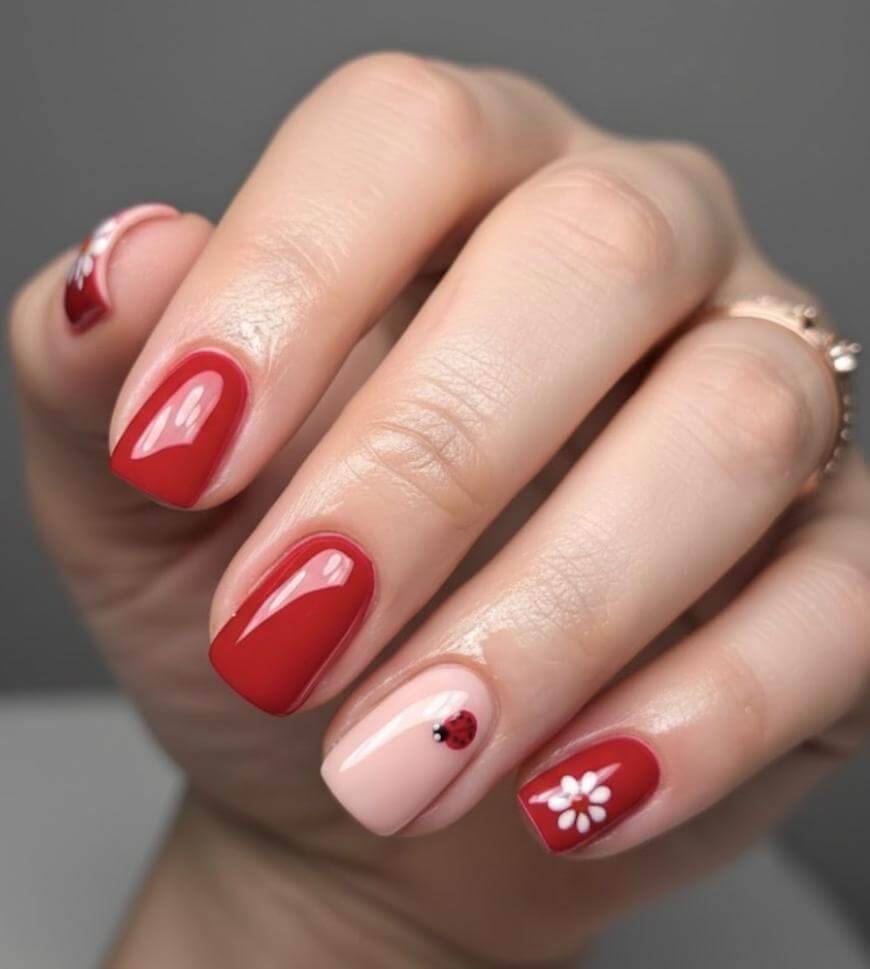 A hand with manicured nails featuring red polish, a light pink accent nail with a small ladybug, and another accent nail with a white daisy design. The nails are short and square-shaped.