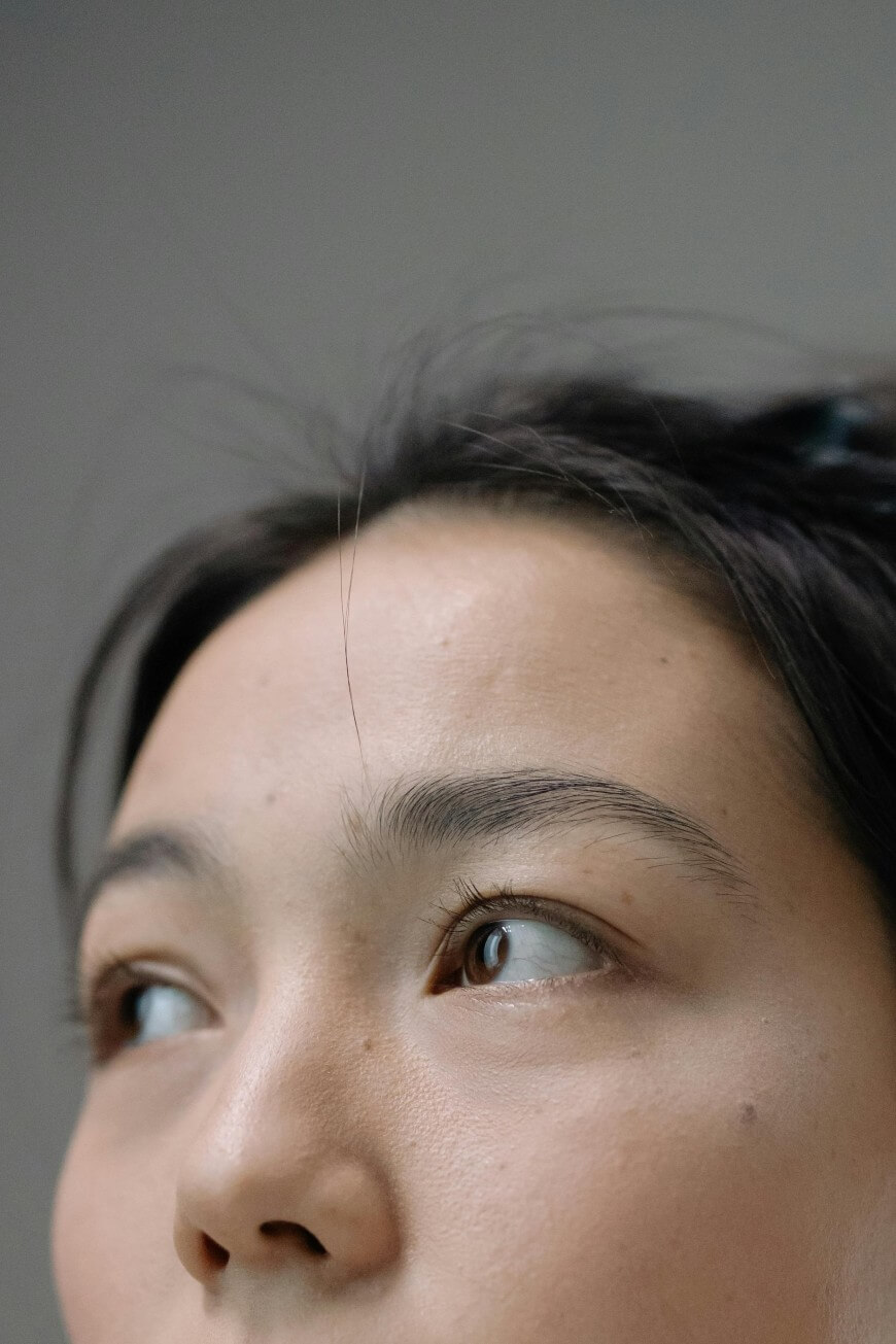 Close-up of a persons face, focusing on the forehead, eyes, and eyebrows. The person is gazing upward to the left, with natural skin texture and loose hair strands visible. The background is neutral and plain.
