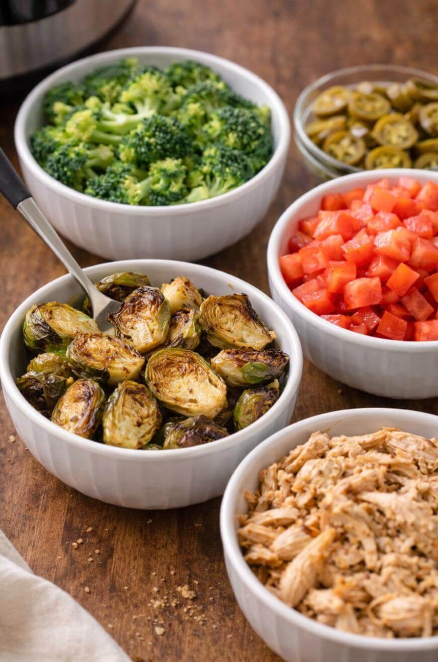 Four white bowls on a wooden table filled with steamed broccoli, roasted Brussels sprouts, diced tomatoes, shredded chicken, and a small bowl of sliced jalapeños in the background.