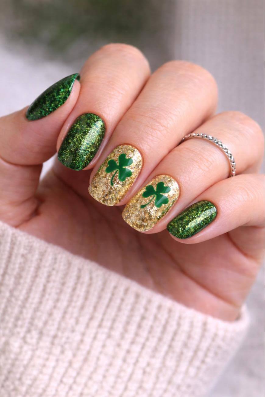 A hand with green glittery nail polish and gold glitter accent nails featuring green shamrock designs, wearing a silver ring and a beige sweater.