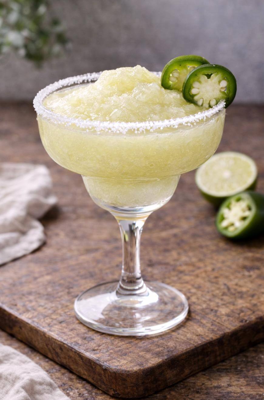 A frozen margarita in a glass with a salted rim, garnished with two slices of jalapeño. A halved jalapeño and lime are in the background on a wooden surface.