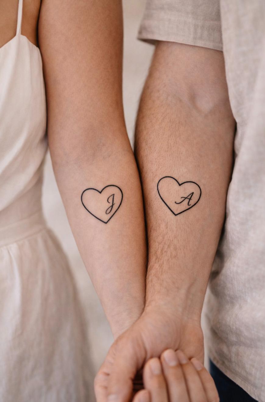 Two people standing side by side show matching heart outline tattoos on their inner forearms; one tattoo has the letter “J” and the other has the letter “A” inside the heart shapes.