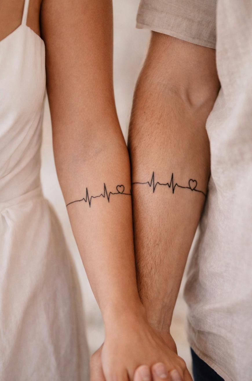 Two people standing side by side with matching forearm tattoos featuring a heartbeat line and a small heart. They are holding hands, and both are wearing light-colored clothing.