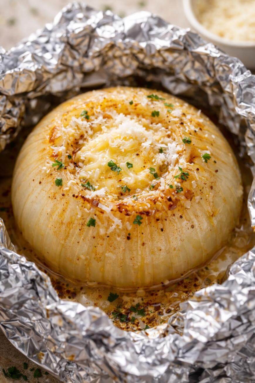 A whole baked onion topped with melted cheese, herbs, and spices, sitting on crinkled aluminum foil. The onion looks soft and golden, with a sprinkle of chopped parsley and grated cheese on top.