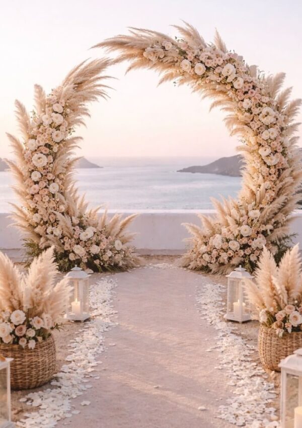 A circular floral arch decorated with pampas grass and white flowers stands on a terrace overlooking the sea. White rose petals line the aisle, with lanterns and woven baskets of pampas grass set along the path.