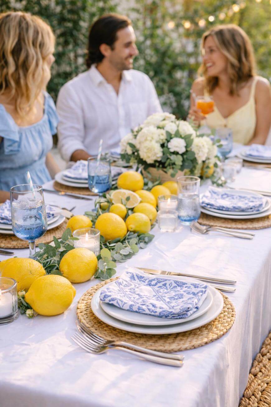 Three people sit at an outdoor table decorated with lemons, greenery, blue and white plates, and woven placemats. They are smiling and talking, with candles and drinks on the table.