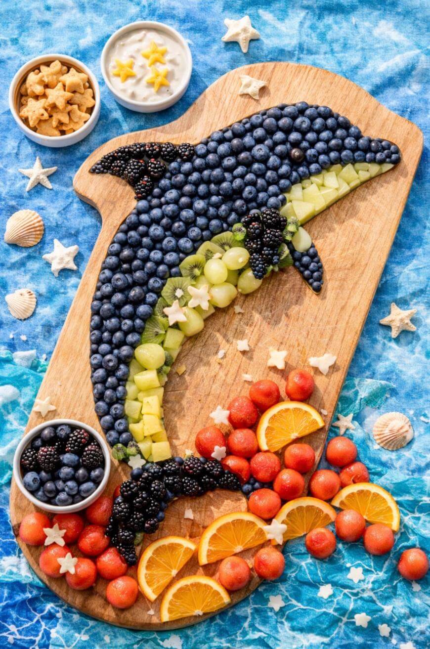 A wooden board displays a dolphin shape made of blueberries, blackberries, grapes, and honeydew, with orange slices, watermelon balls, and yogurt cups arranged around it on a blue ocean-themed background.
