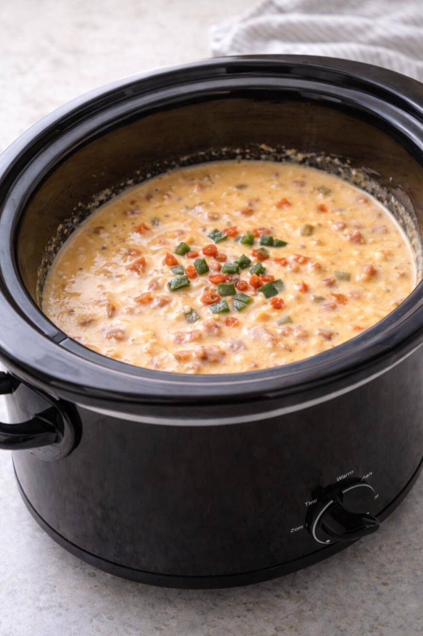 A black slow cooker filled with creamy, chunky cheese dip topped with chopped green herbs and small bits of tomato or pepper, sitting on a light-colored surface.