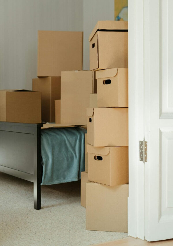 Several cardboard moving boxes are stacked and scattered in a sunlit room next to a black bed frame with a green blanket, partially visible through an open white door.