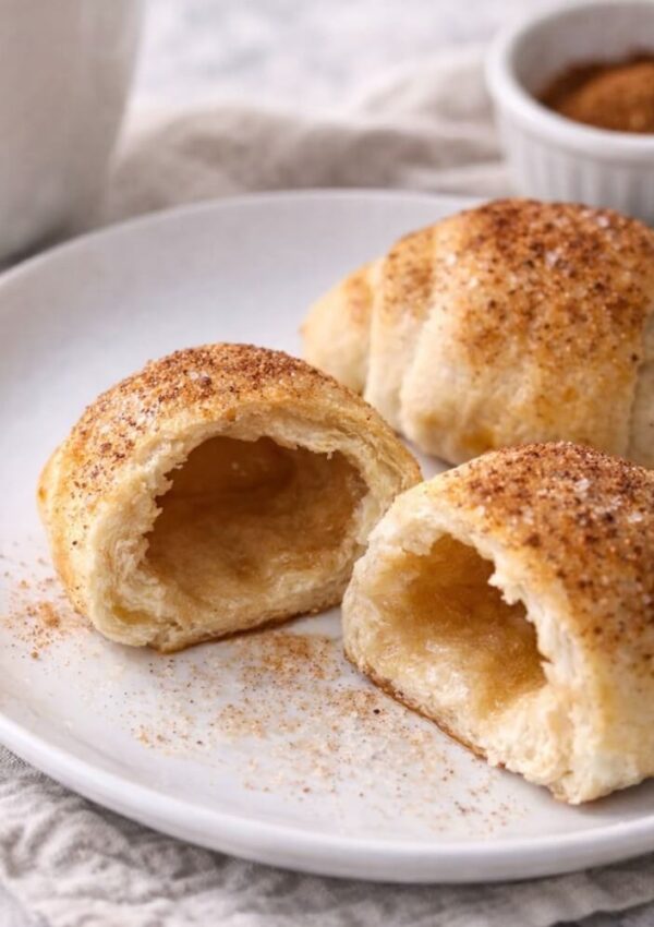 A plate with flaky, golden-brown crescent rolls dusted with cinnamon sugar. One roll is cut open, showing a hollow, airy inside. A small bowl with more cinnamon mixture is in the background.
