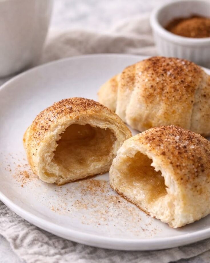 A plate with flaky, golden-brown crescent rolls dusted with cinnamon sugar. One roll is cut open, showing a hollow, airy inside. A small bowl with more cinnamon mixture is in the background.
