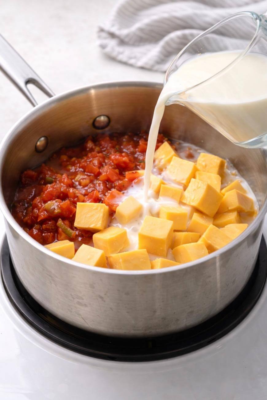 A saucepan on a stovetop contains diced tomatoes, cubes of processed cheese, and is having milk poured in from a glass measuring cup; a light cloth is in the background.