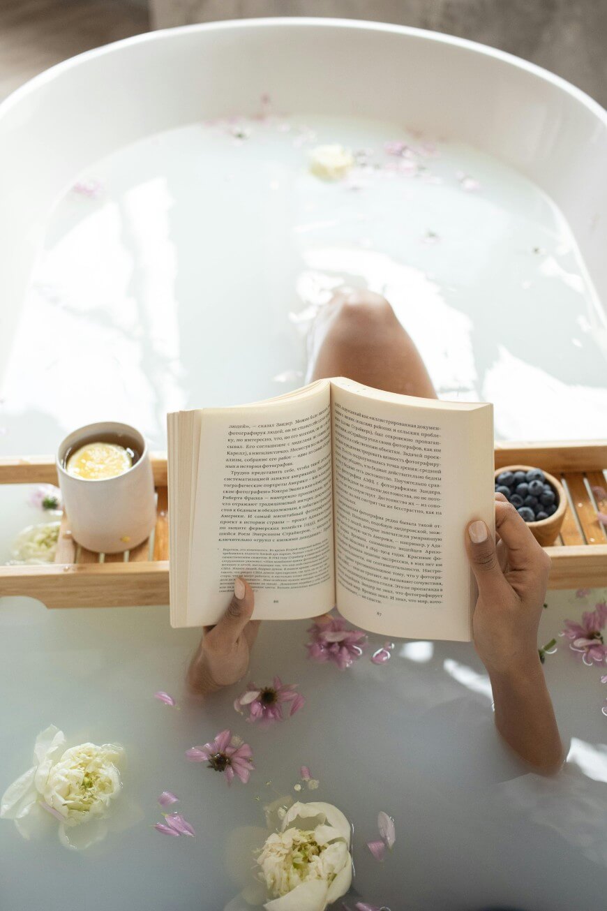 A person relaxes in a milky bath with flower petals, reading a book. A wooden tray holds a cup of coffee and a bowl of blueberries above the tub. Only their hands and knees are visible.