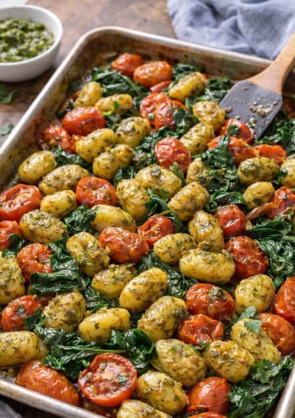 A sheet pan filled with roasted gnocchi, cherry tomatoes, and wilted spinach, all coated with green pesto. A wooden spatula and a small bowl of extra pesto are nearby.