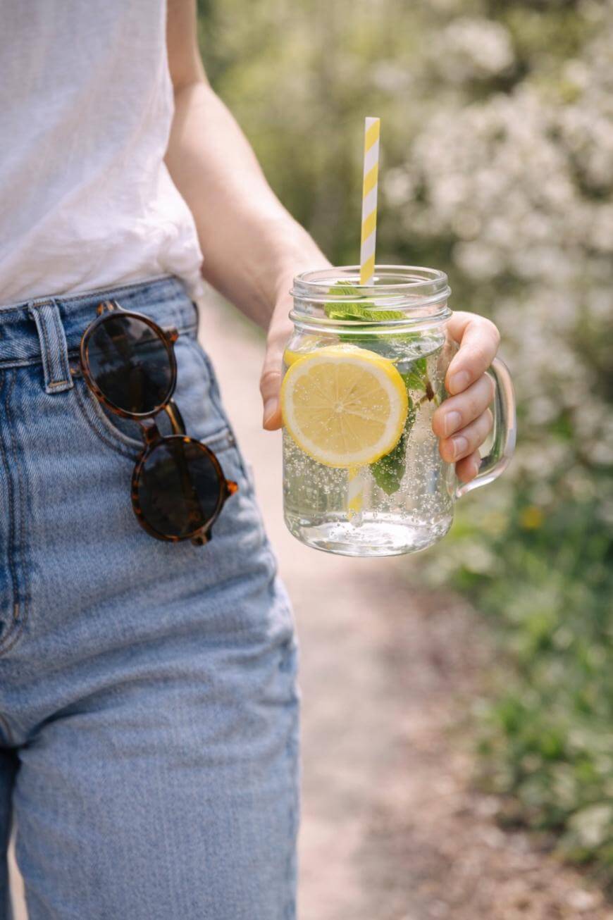 A person in blue jeans and a white shirt holds a mason jar of sparkling water with lemon, mint, and a striped straw. Sunglasses are hooked on their pocket; greenery is blurred in the background.