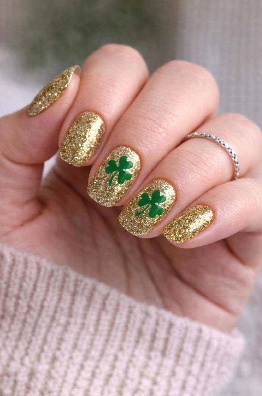 A hand with gold glittery nail polish, featuring green shamrock designs on the middle and ring fingernails. The person wears a silver ring and a beige knit sleeve is visible.