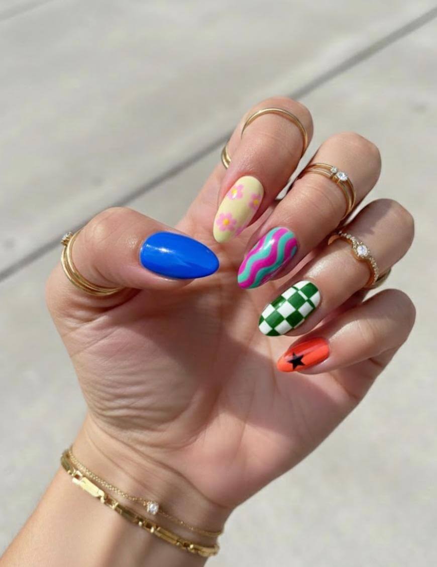 A hand with long, almond-shaped nails, each featuring a unique design: solid blue, pastel yellow with pink flowers, pink and green wavy lines, green and white checkerboard, and orange with a small black star.