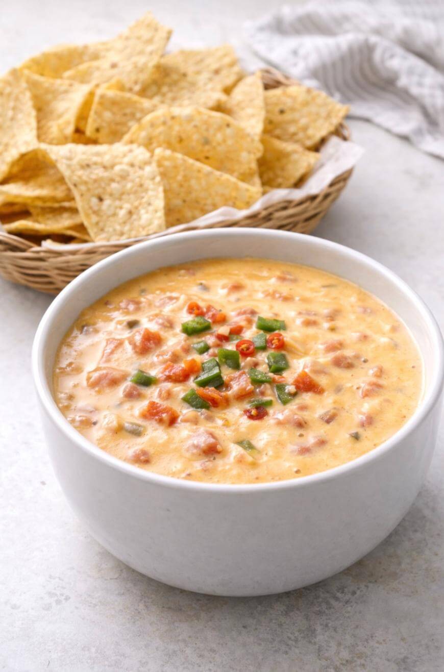 A white bowl filled with creamy queso dip topped with chopped green and red peppers, sits in front of a basket of crispy tortilla chips on a light surface.