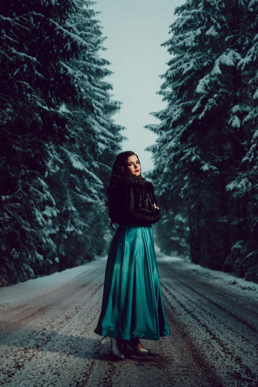 A woman in a long teal dress and dark fur shawl stands on a snowy forest road, surrounded by tall, snow-covered trees under a cloudy sky.