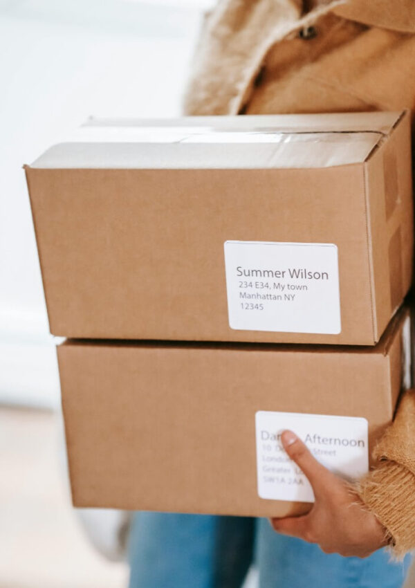 A person wearing a tan jacket holds two brown cardboard boxes with address labels; the top label reads Summer Wilson, 234 E84, My town, Manhattan NY 12345.