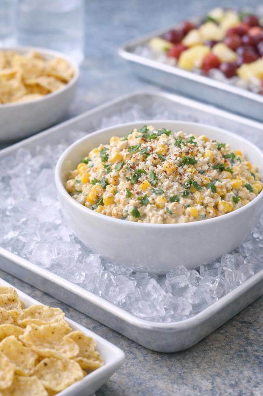 A white bowl filled with creamy corn dip garnished with herbs, sitting on a bed of ice, with tortilla chips and a tray of fruit in the background.