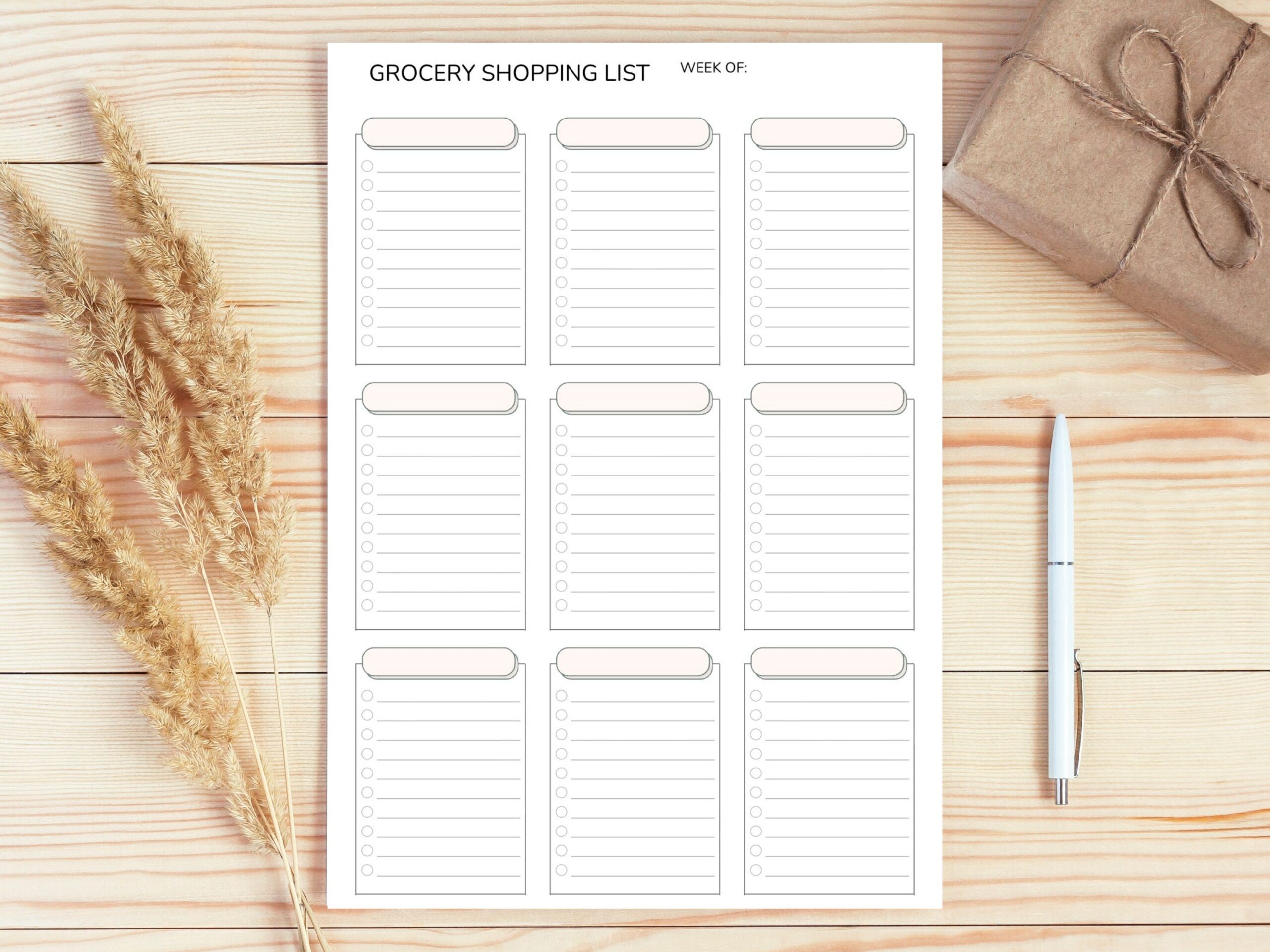 The Minimalist Grocery Shopping List Printable PDF – Categorised Weekly Checklist (A4, A5, Letter & Half Size) lies on a wooden surface with a pen on the right, dried grasses on the left, and a wrapped gift in the top right corner.