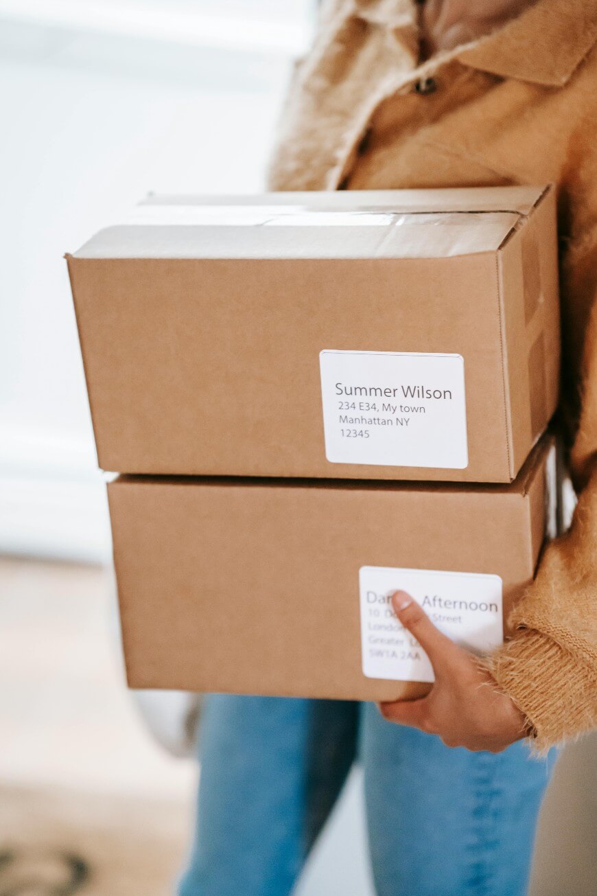 A person wearing a tan jacket holds two brown cardboard boxes with white address labels; the top label reads Summer Wilson. The boxes are being carried indoors.