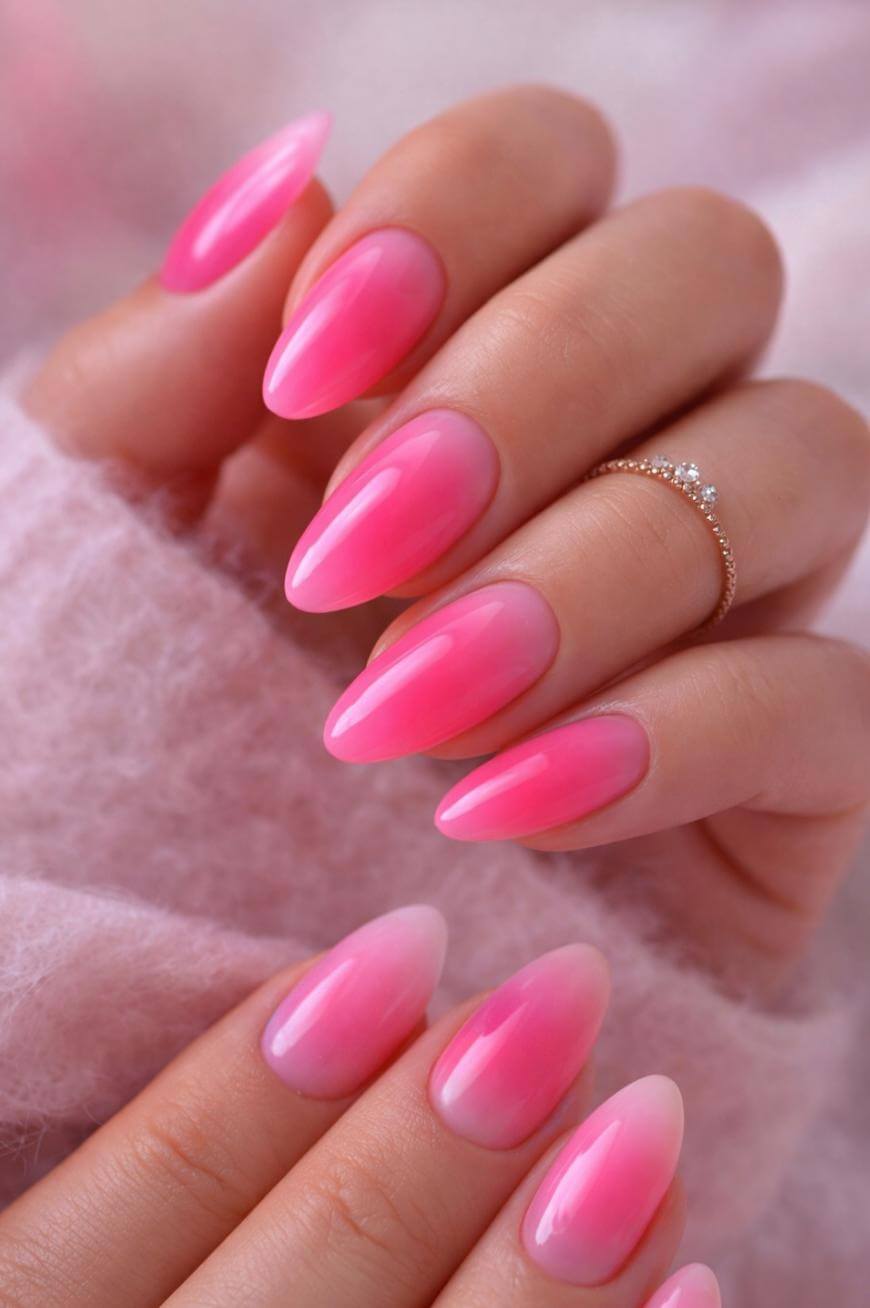 A close-up of hands with long, almond-shaped nails painted in a glossy pink ombre style. One hand has a delicate, gold ring with small diamonds. The background is soft and fuzzy, in light pink tones.