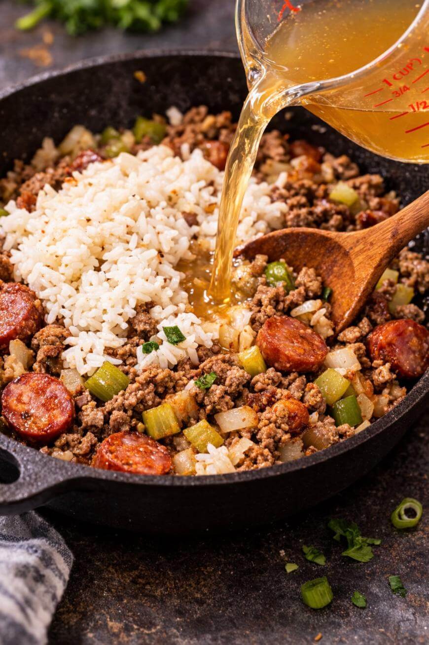 A close-up of a skillet with cooked ground meat, sliced sausage, chopped celery, onions, and white rice, as broth is poured in from a measuring cup. A wooden spoon rests in the skillet.