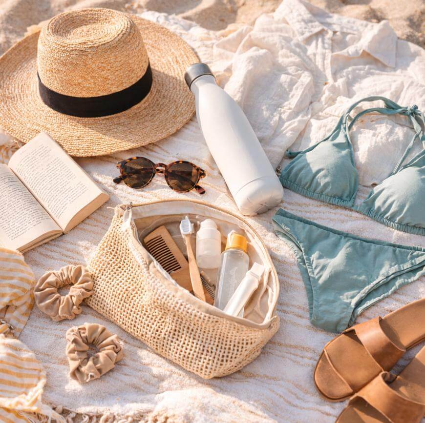 A flat lay of beach essentials on a striped towel, including a straw hat, sunglasses, a book, a bikini, a reusable water bottle, sandals, scrunchies, and a woven bag filled with toiletries and a comb.