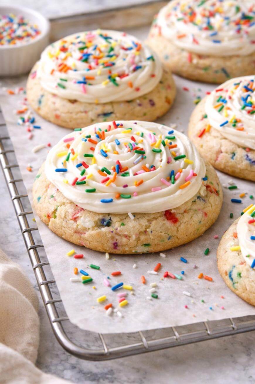 Large sugar cookies topped with swirled white frosting and colorful rainbow sprinkles are arranged on a parchment-lined cooling rack. More sprinkles are scattered around the cookies.