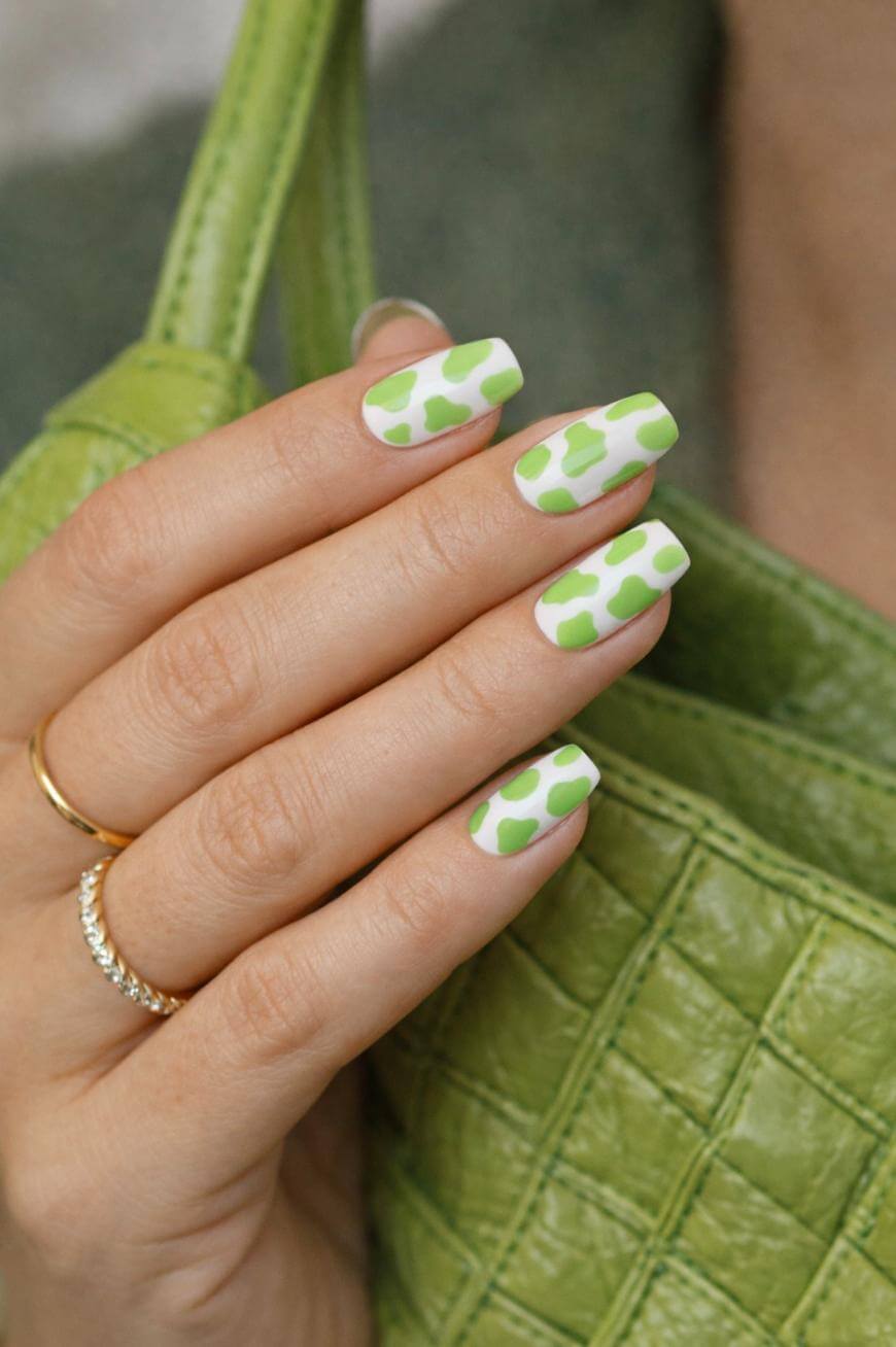A hand with long, square-shaped nails featuring a white base and green abstract spots, holding a light green textured handbag. The person wears two gold rings.