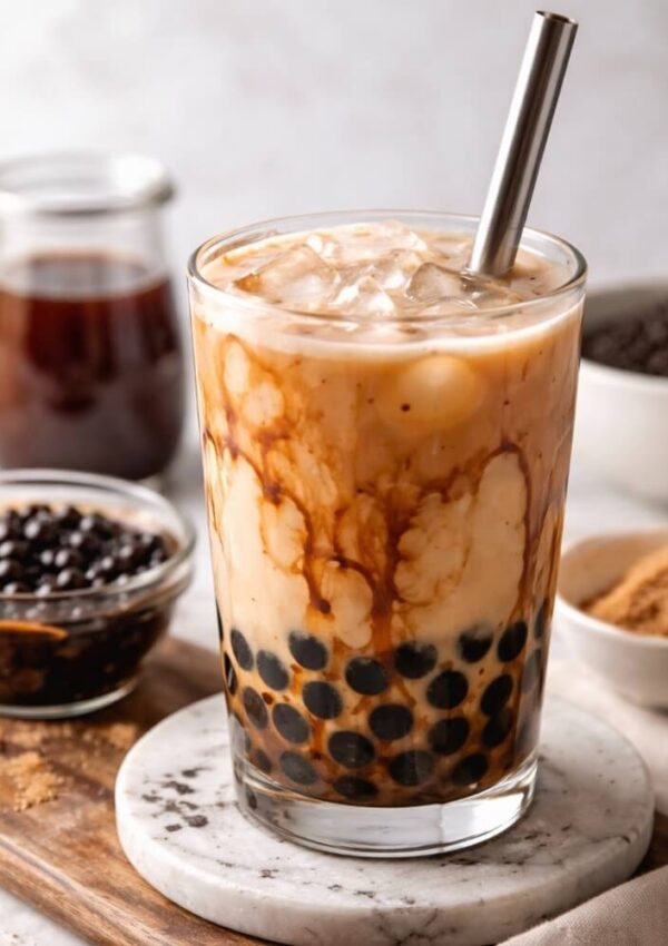 A glass of iced bubble tea with dark tapioca pearls, swirls of milk, and a metal straw, surrounded by ingredients like brown sugar, black pearls, and syrup on a light background.