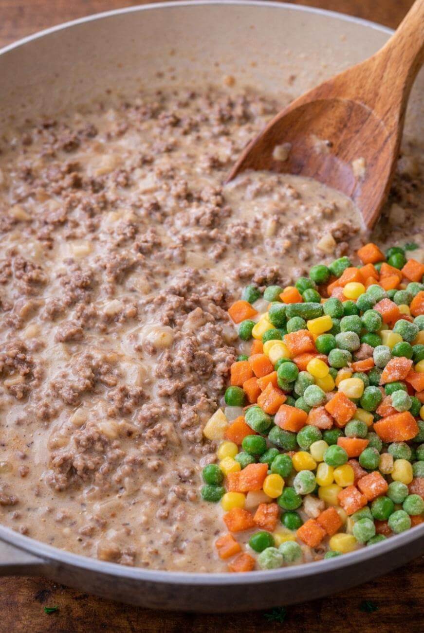 A skillet with creamy ground beef mixture on one side and a pile of frozen mixed vegetables (corn, peas, carrots, green beans) on the other, with a wooden spoon resting in the pan.
