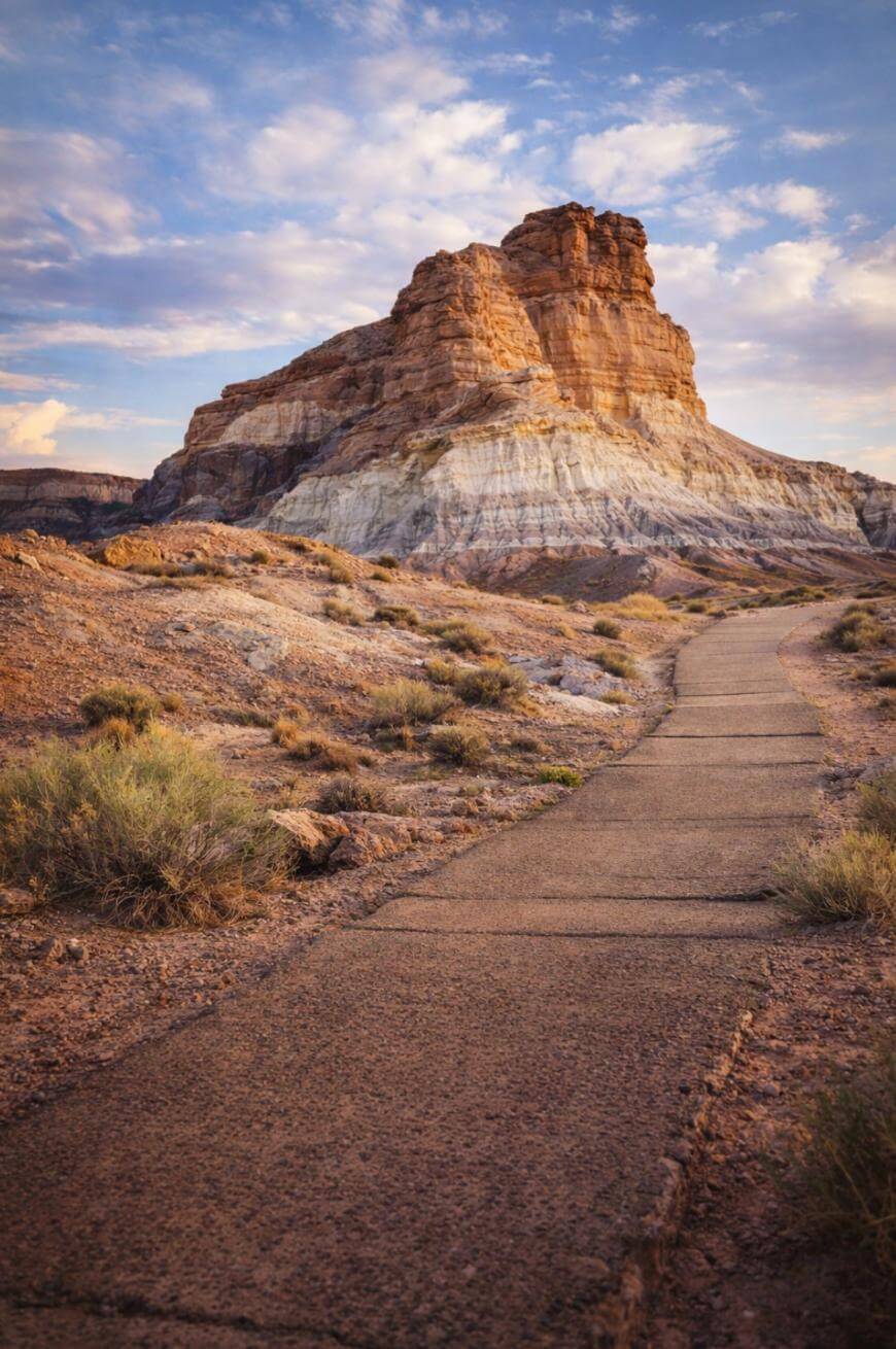 A paved path winds through dry desert terrain toward a large, layered rock formation under a partly cloudy sky at sunset.
