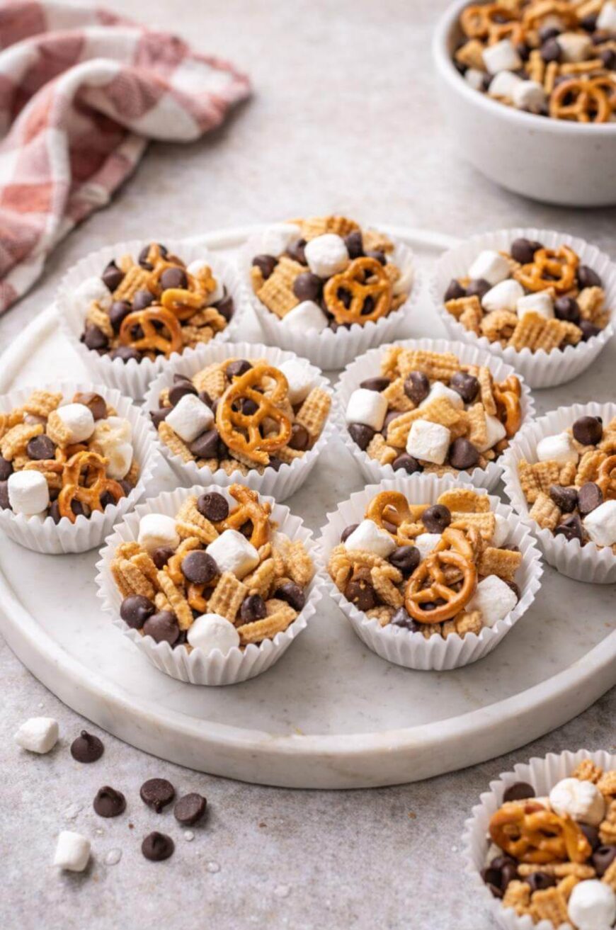 Mini snack mix cups filled with pretzels, chocolate chips, cereal squares, and mini marshmallows are arranged on a white plate, with a bowl of extra mix and a red checkered cloth in the background.